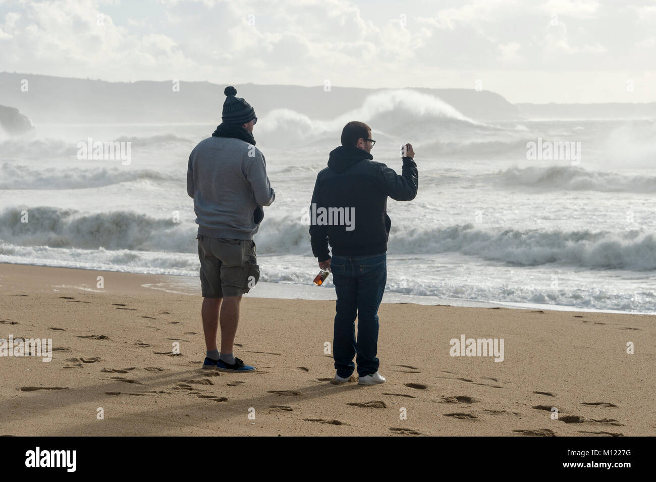 two guys watching the big waves at Nazare, Portugal. Taking a photo on a smart phone. Stock Photo