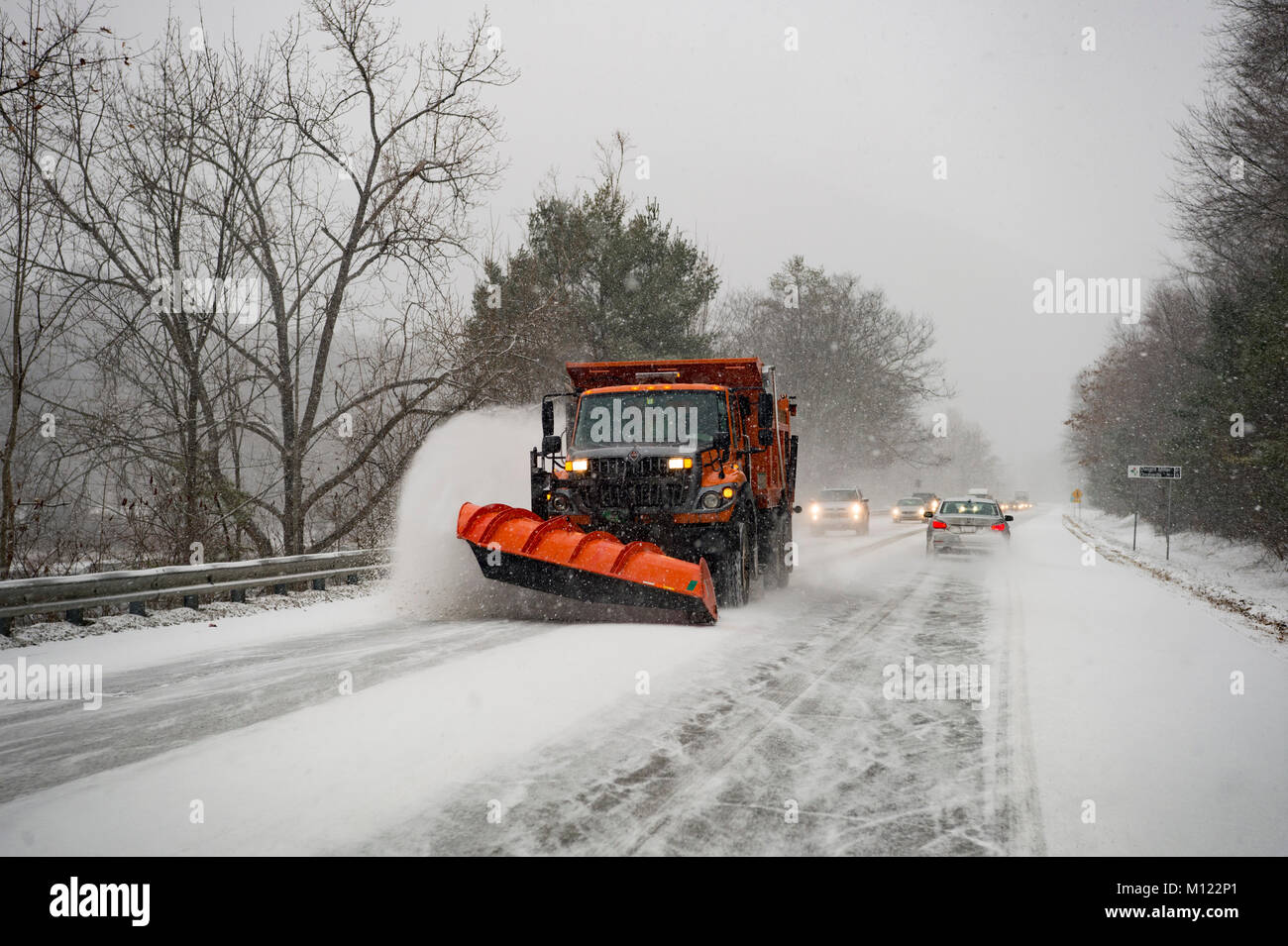 A snow plow clears the road followed by a line of traffic as it snows