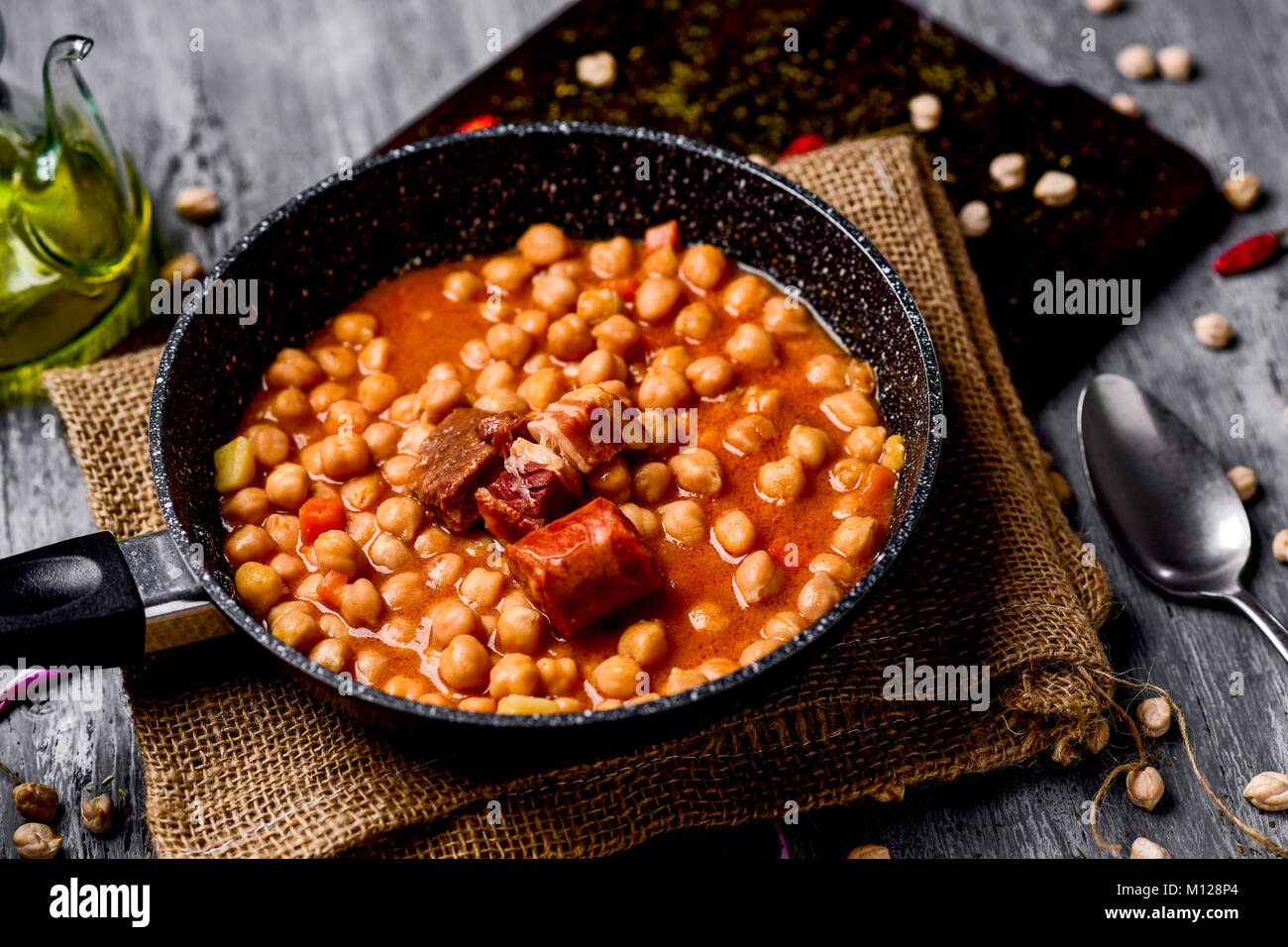 a stone frypan with garbanzos a la riojana, a spanish chickpeas stew, on a wooden chopping board placed on a rustic wooden table next to a cruet with  Stock Photo