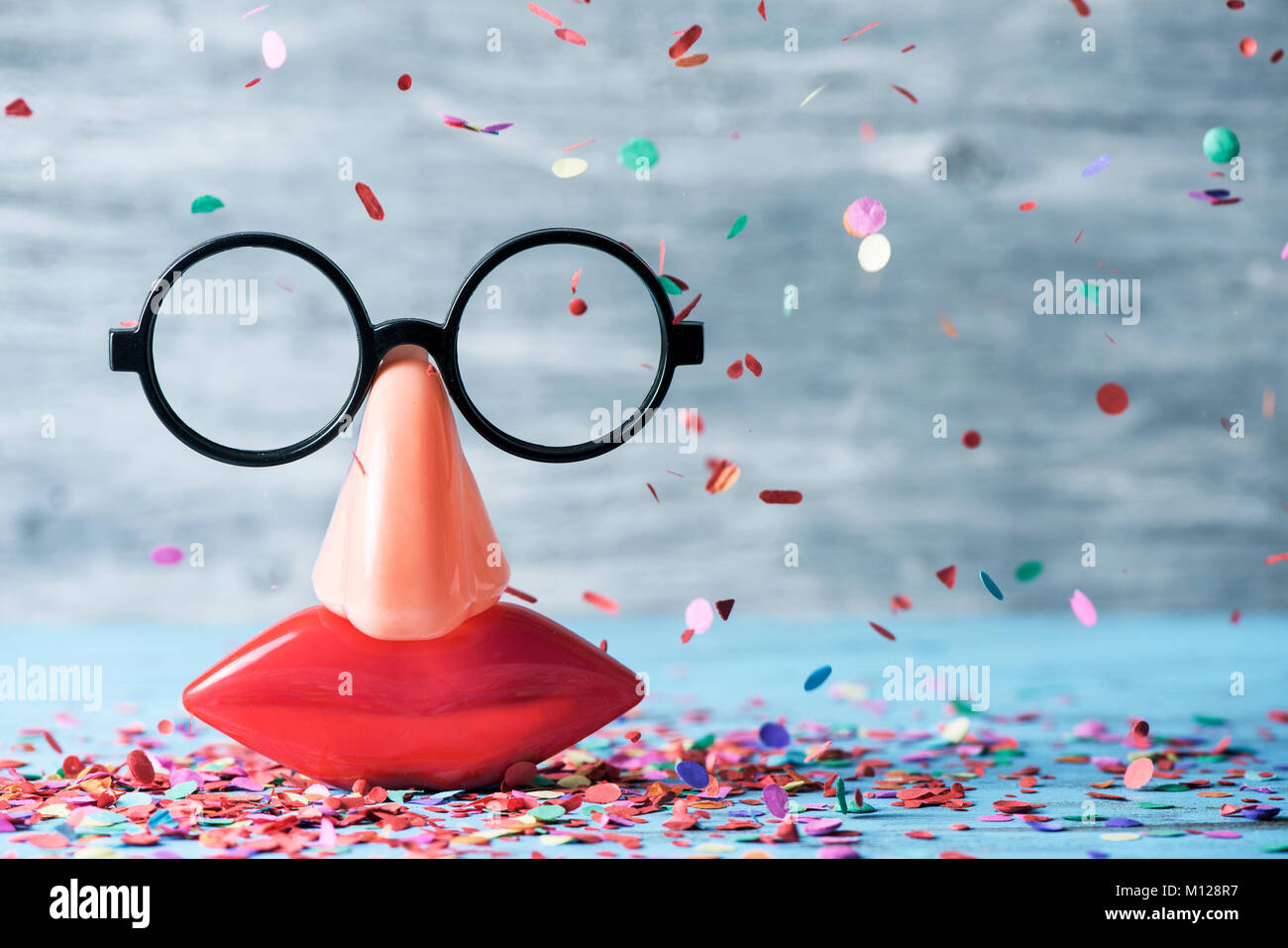 closeup of a pair of fake black glasses, a nose and a mouth forming the face of a person on a blue rustic wooden surface full of confetti Stock Photo