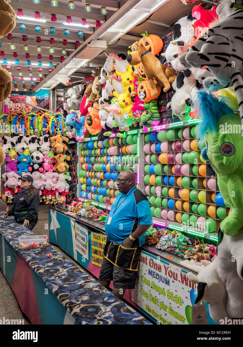 Carnival booth workers one male one female in a dart throwing carnival game with colorful balloons, cupie dolls and stuffed animals for prizes. Stock Photo