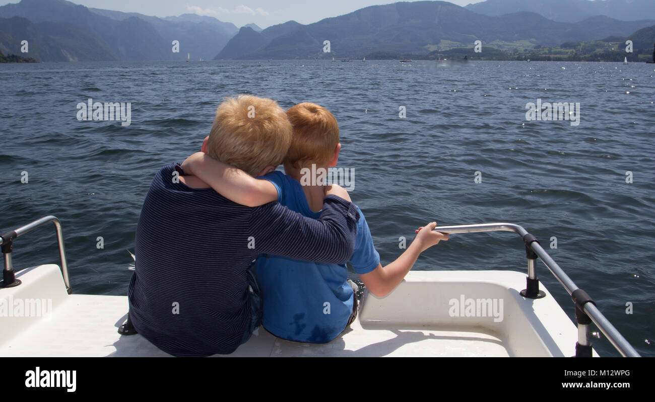 Two young boys sitting arm in arm on the deck of a white boat looking out over the sea to distant mountains on the coastline in a rear view Stock Photo