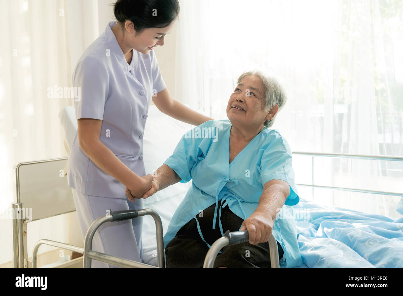 Asian young nurse supporting elderly patient disabled woman in using walker in hospital. Elderly patient care concept. Stock Photo