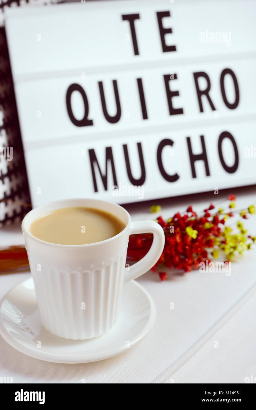 closeup of a white ceramic cup with coffee on a table, a bunch of flowers and a lightbox in the background with the text te quiero mucho, I love you s Stock Photo