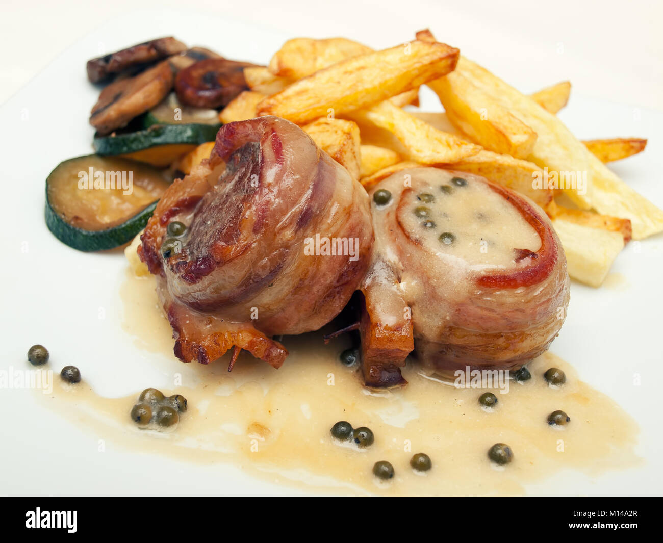 Closeup view of tasty bacon wrapped steaks with green pepper sauce, french fry potatoes and roasted vegetables. Stock Photo