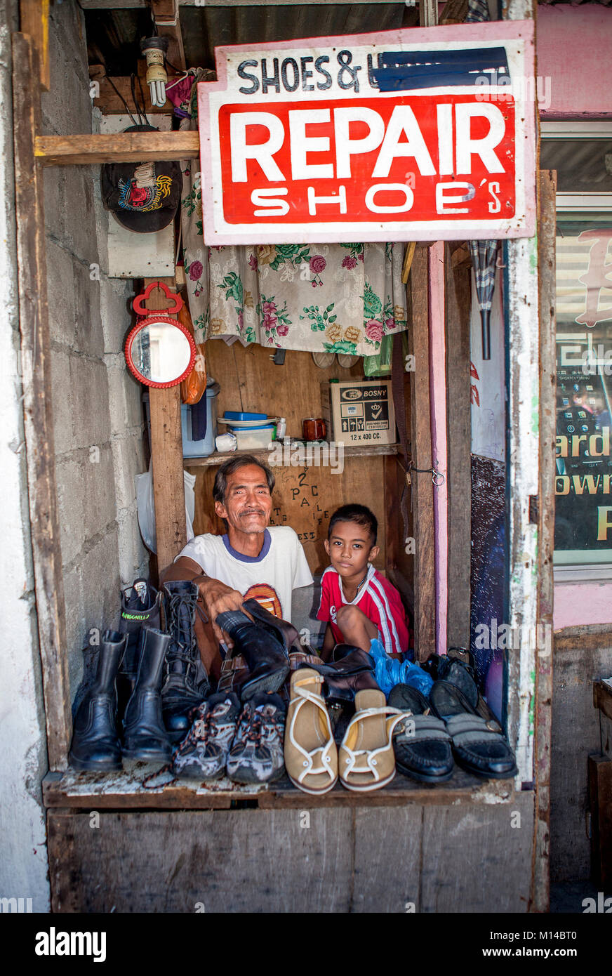 A small, family-run cobbler shop tended by a grandfather with his grandson is only a few feet wide in Barretto, Luzon, Philippines. Stock Photo