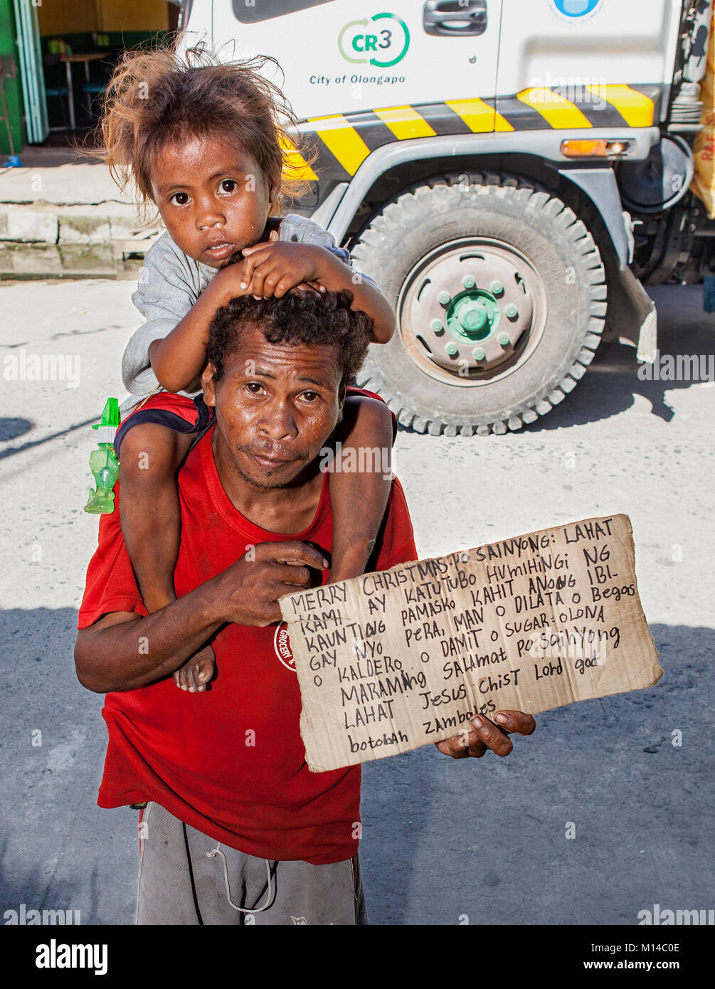 A Filipino Aeta man carries his child on his shoulders and a sign asking for food and work and with a Christmas wish in Barretto Town, Luzon, Philippi. Stock Photo
