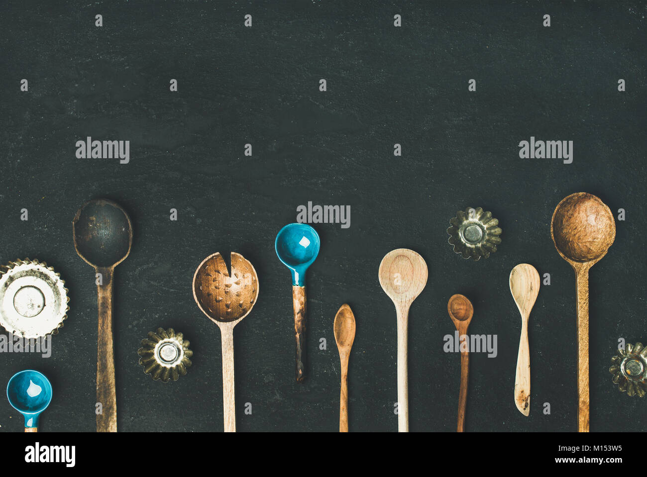 Flat-lay of various old vintage kitchen spoons and baking tin molds over black stone background, top view, copy space, horizontal composition. Rustic  Stock Photo