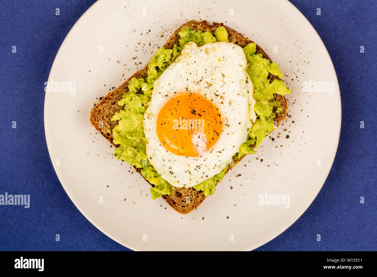 Fried Egg Sunny Side Up on Crushed Avocado And Rye Bread Open Faced Sandwich Against A Blue Background Stock Photo