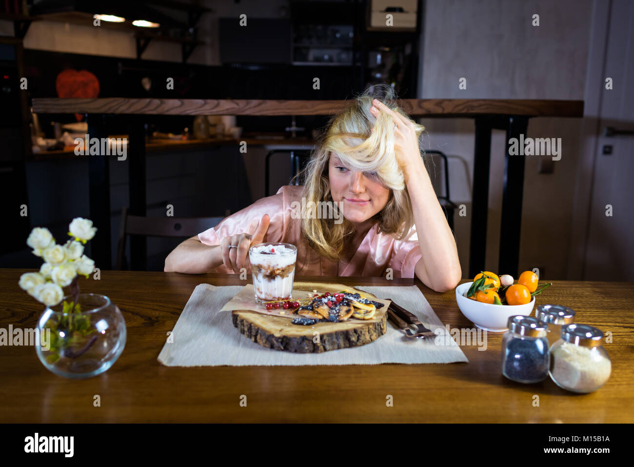 Funny blonde sits at the table with food against modern kitchen in the morning. Young woman eats fresh pancakes with dessert on the breakfast Stock Photo