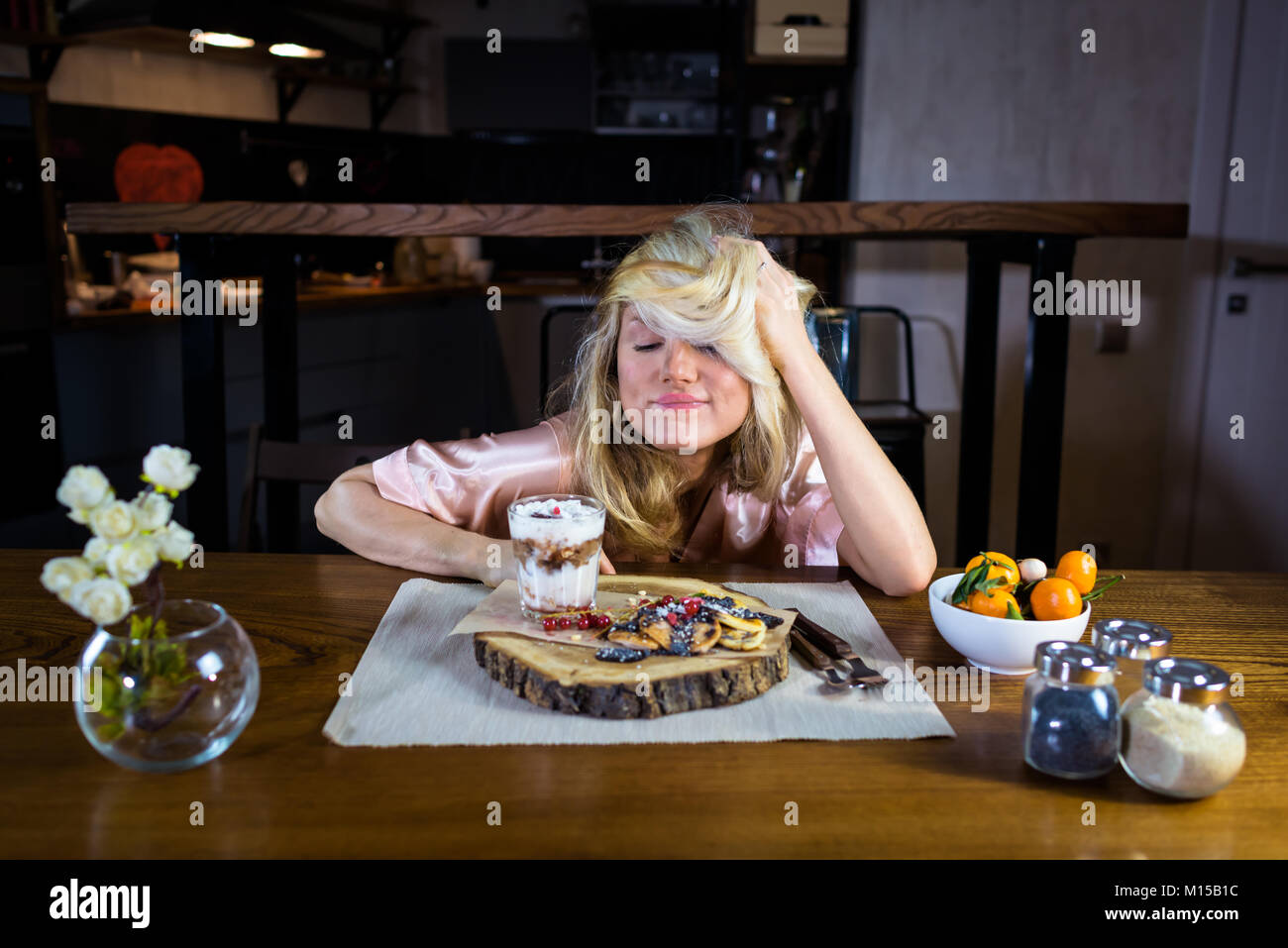 Young woman eats fresh pancakes on the breakfast at home alone. Funny blonde sits at the table with food against modern kitchen in the morning Stock Photo