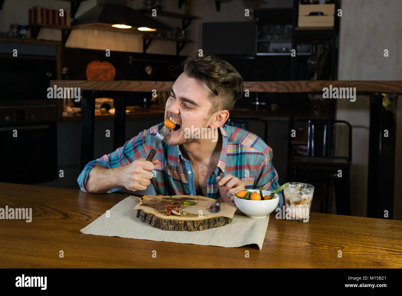 Hungry person emotionally eats food on the breakfast. Young man sits at the table in the modern home kitchen. Beautiful guy early in the morning Stock Photo