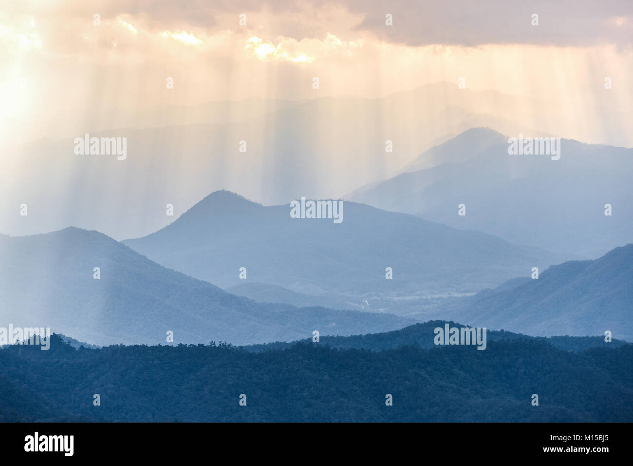 Sunbeam from cloudy sky shining through fog to blue complex mountains range in morning after very cold night of winter season. Stock Photo