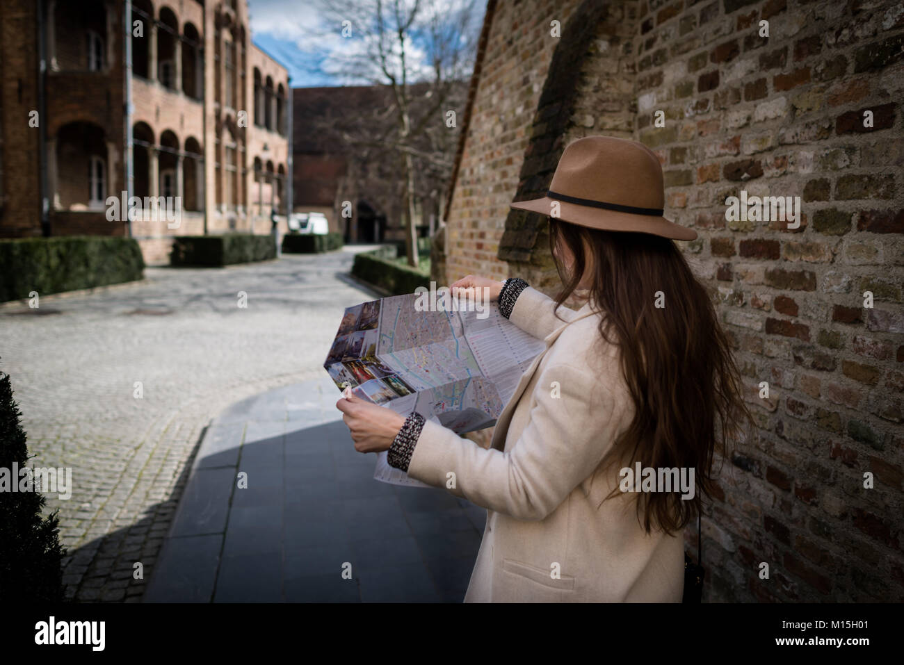 Elegant female tourist exploring map while standing in alley near building. Young woman traveler reading geographical atlas while touring in old town Stock Photo
