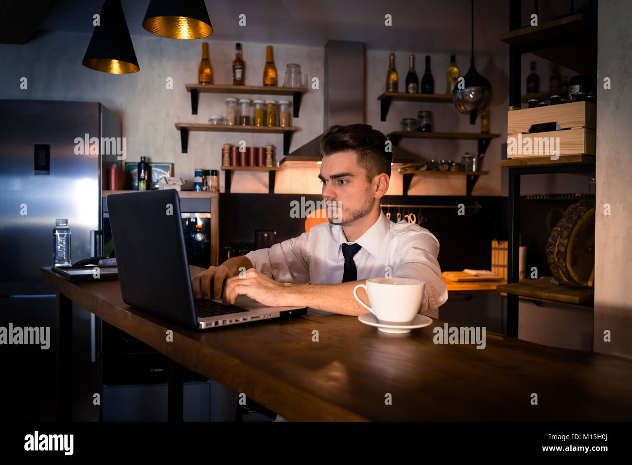 Young man sits in the kitchen at the bar counter and works in laptop. Home atmosphere, work in the evening with cup of coffee. Home office. Stock Photo