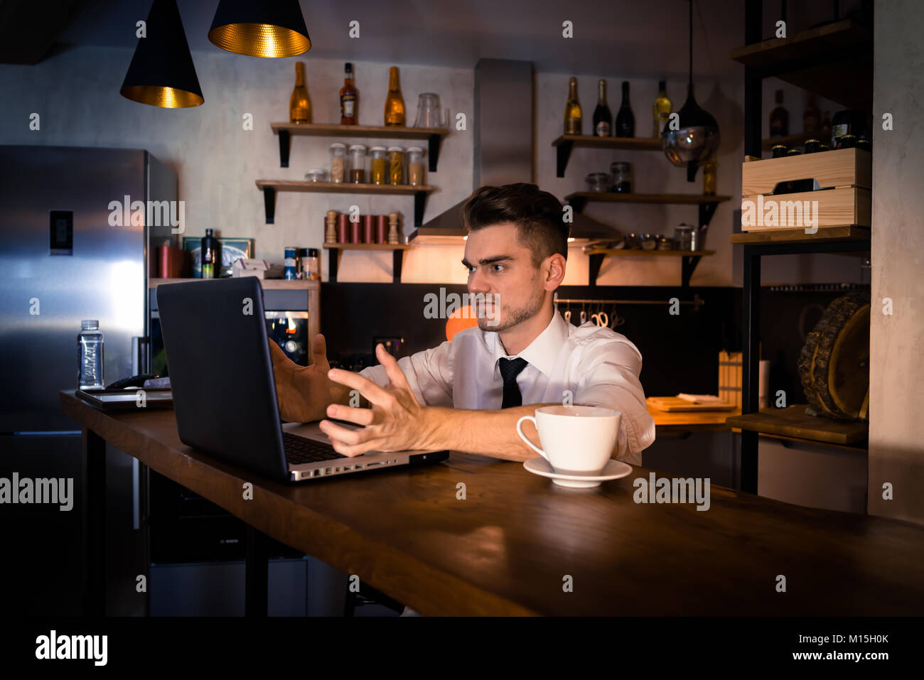 Dissatisfied young man sits in the kitchen at the bar counter and works in laptop. Home atmosphere, work in the evening with cup of coffee Stock Photo