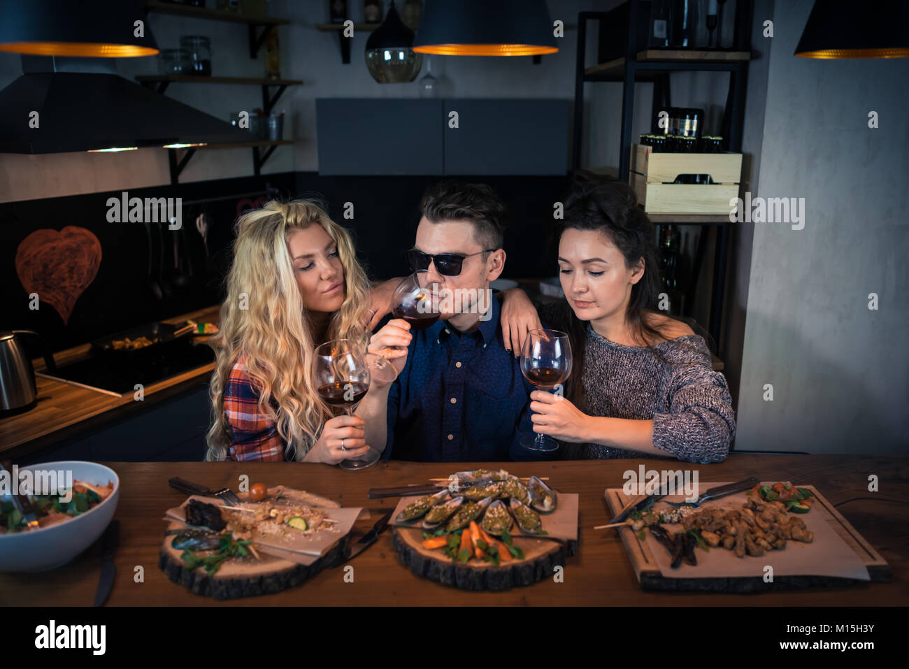 Two young women with a man in sunglass has late dinner in modern home kitchen. Group of beautiful people sits together and eats food with wine Stock Photo