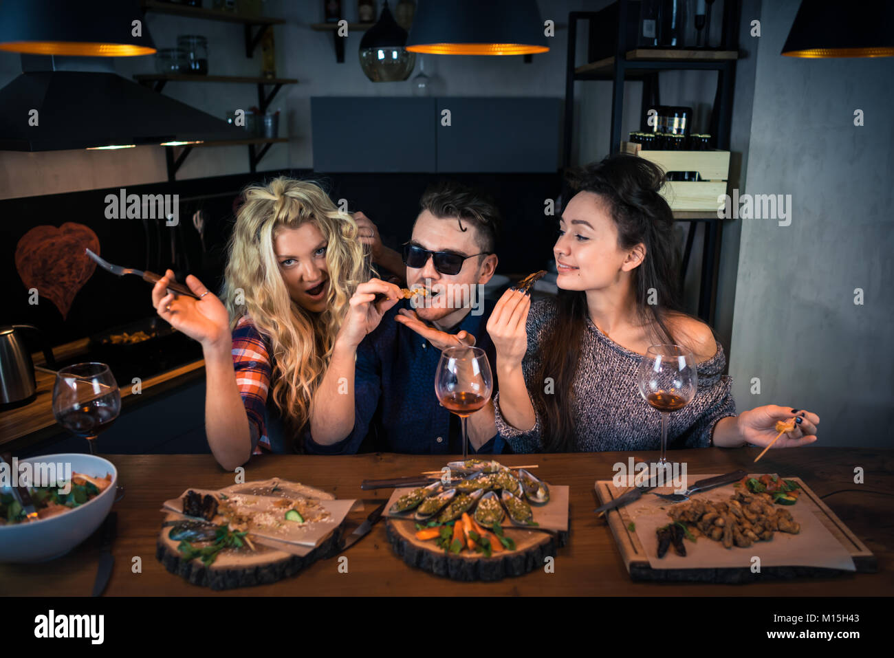 Two young women with a man smiles and has dinner together at the bar counter. Group of beautiful people and eats food with wine and enjoying evening Stock Photo