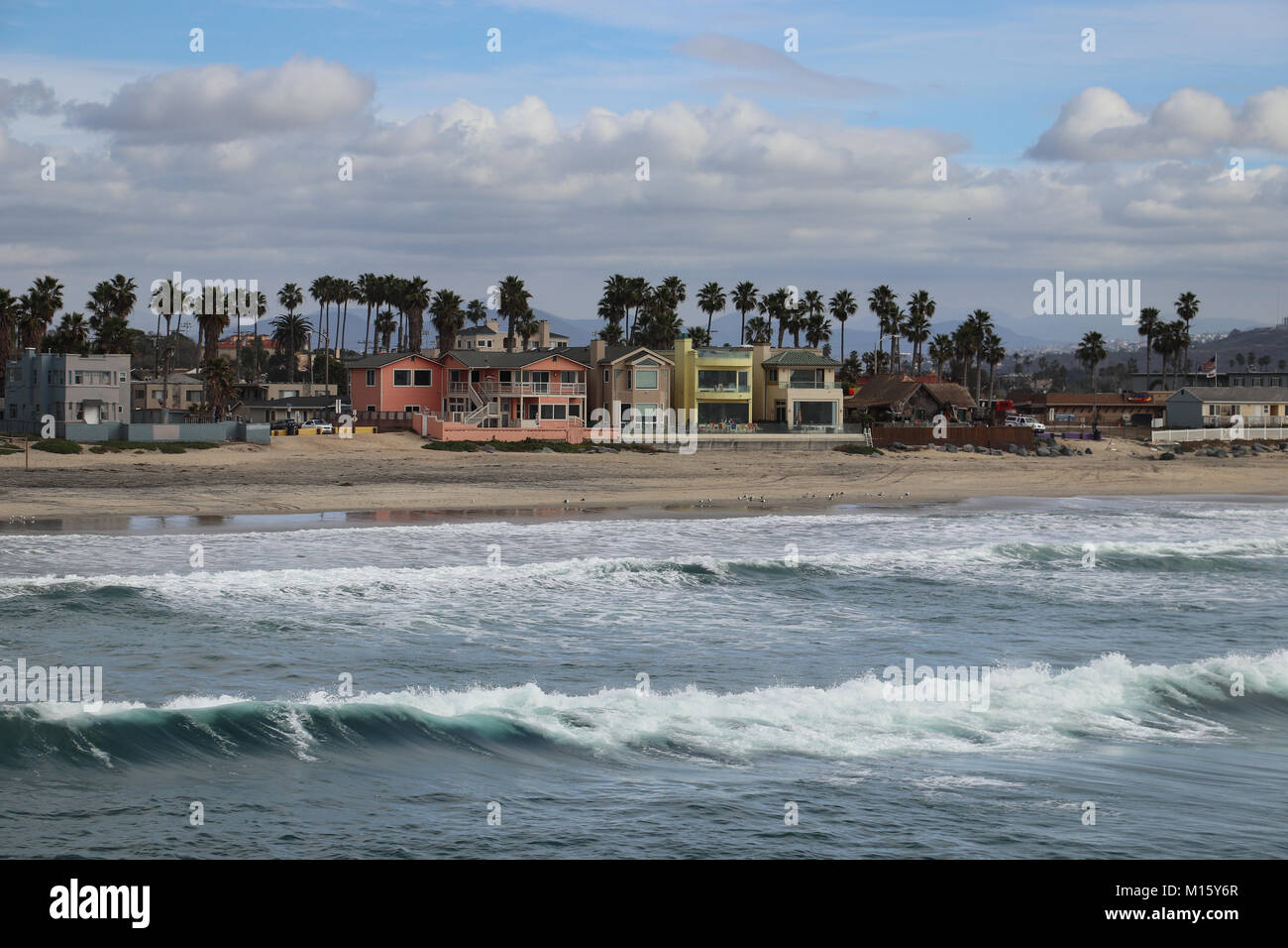 Multicolored beach cottages line the shore at Imperial Beach, California, as seen from the pier.Sets of waves are breaking on the beach, clouds in sky Stock Photo