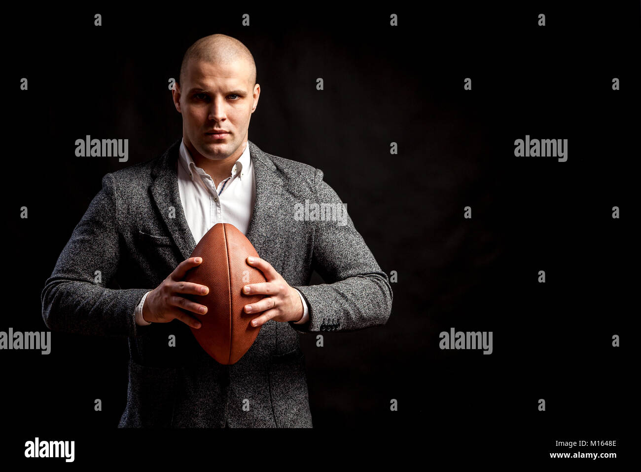 A young bald man in a white shirt, gray suit keeps a rugby ball on a black isolated background Stock Photo