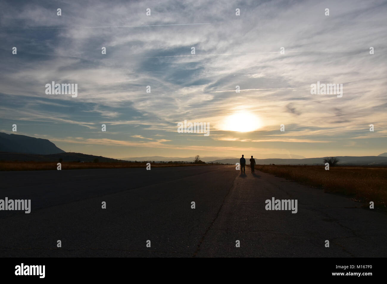 Sunset, flat valley, two people walking on a wide straight empty asphalt road towards the sun at the horizon Stock Photo