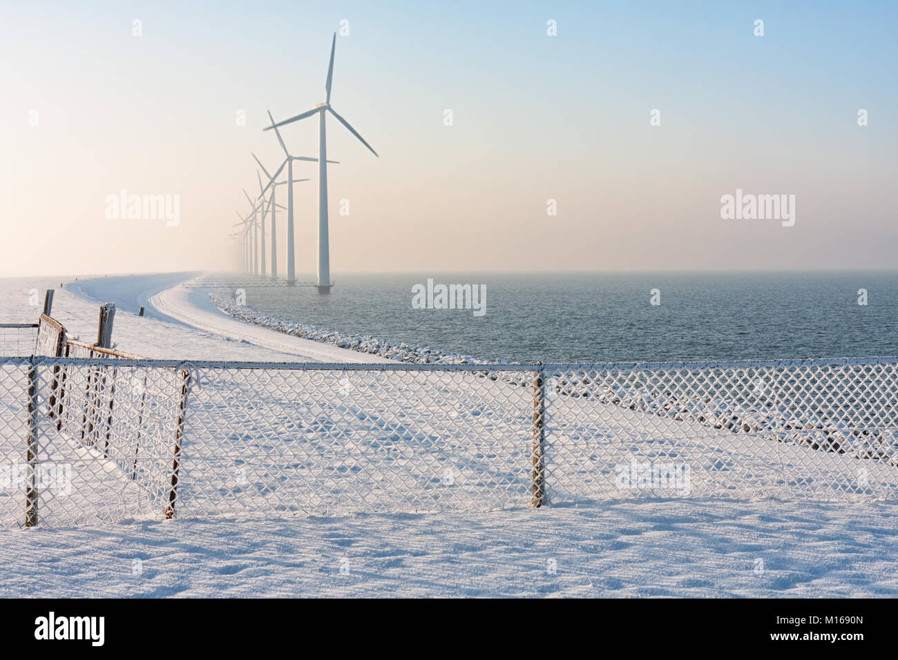 Dutch dike in winter with snow, fence and wind turbines Stock Photo