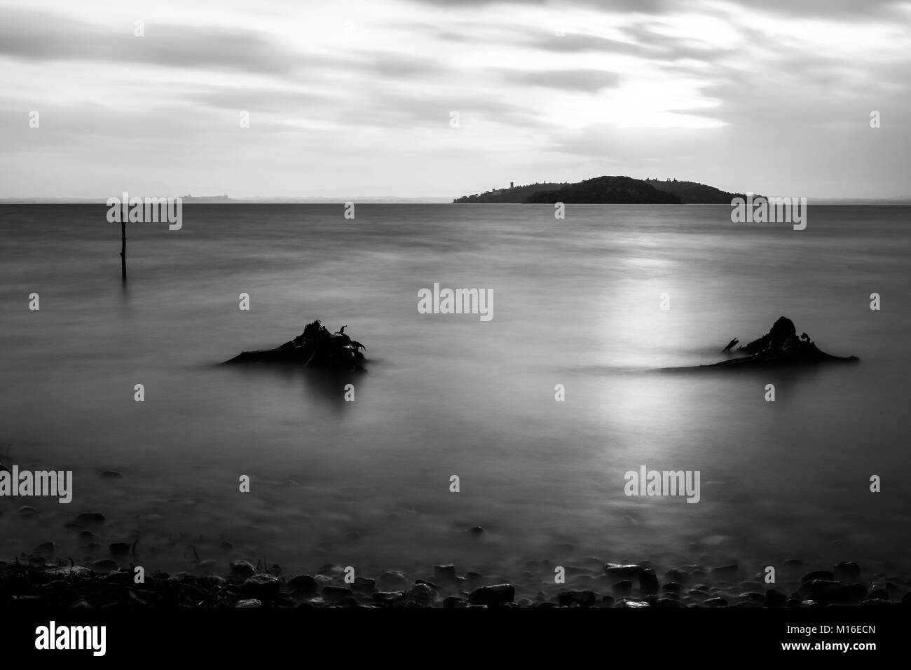 Long exposure view of Trasimeno lake (Umbria) shore, with branches, stones and poles beneath a cloudy sky filtering the sunset Stock Photo