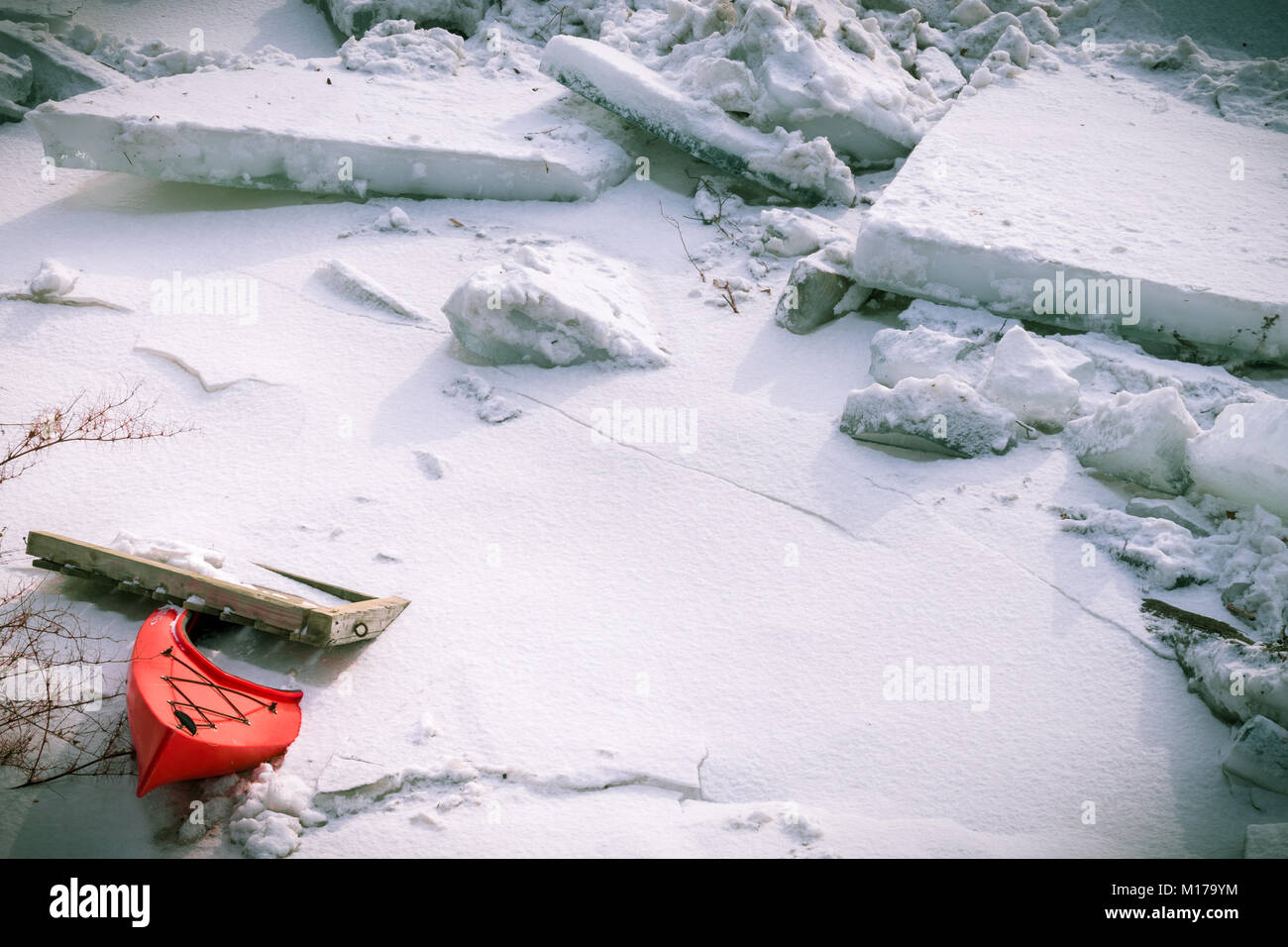 A red kayak and a portion of a wooden dock lay stuck near an ice floe in a frozen river. Stock Photo