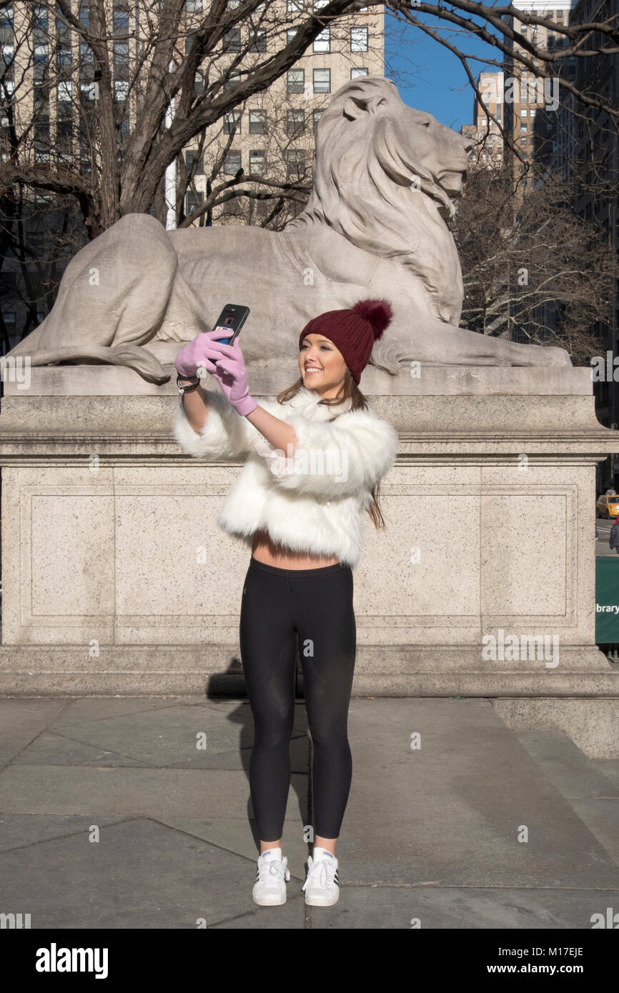 A very pretty young lady takes a selfie in front of the statue of the lion in front of the main branch of the New York Public Library. Stock Photo