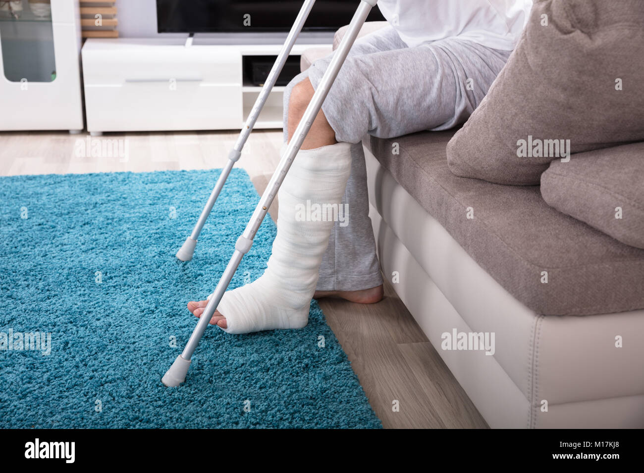 Young Man With Broken Leg Using Crutches To Get Up From Sofa Stock