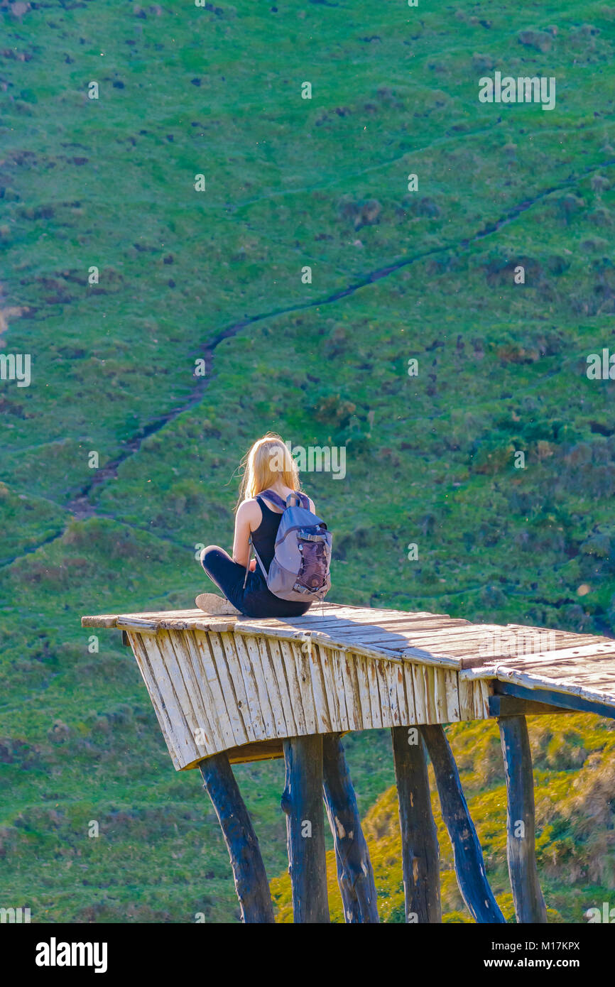Attractive young blonde woman sitting at border of precipice at famous Mirador de las Almas viewpoint, Chiloe Island, Chile Stock Photo