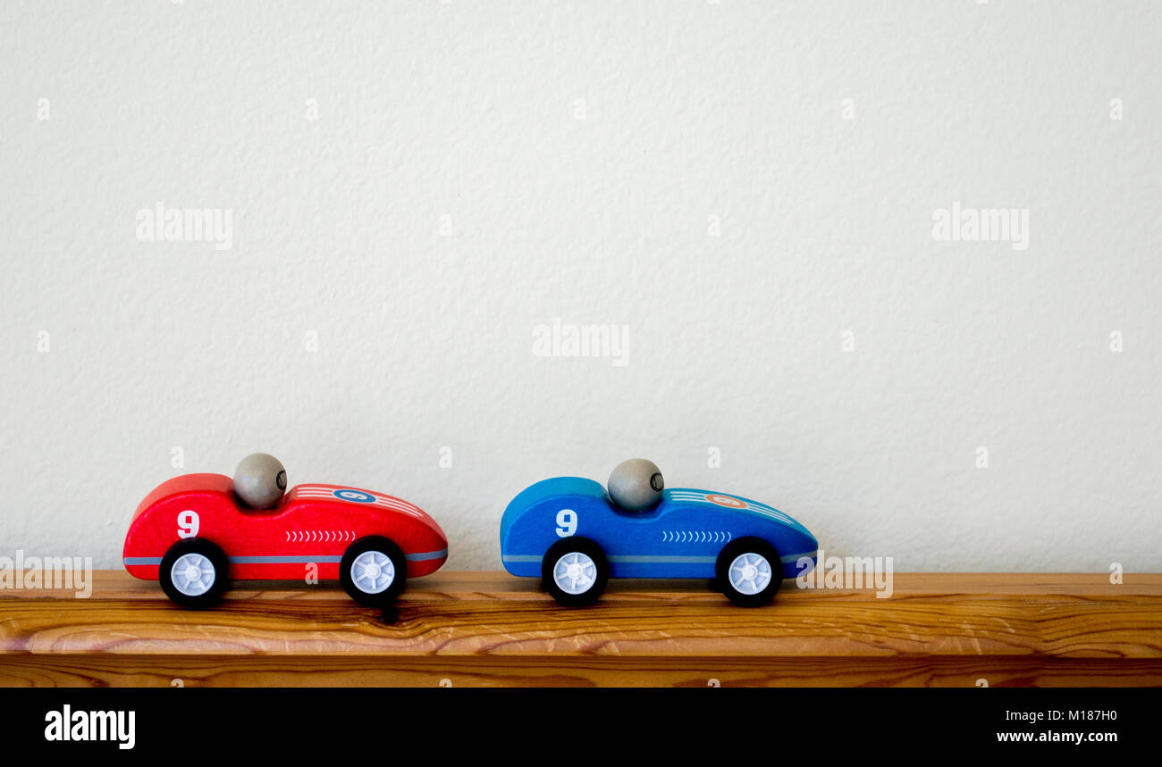 2 colorful wooden toy race cars running along a wood railing against a blank wall with copy space Stock Photo