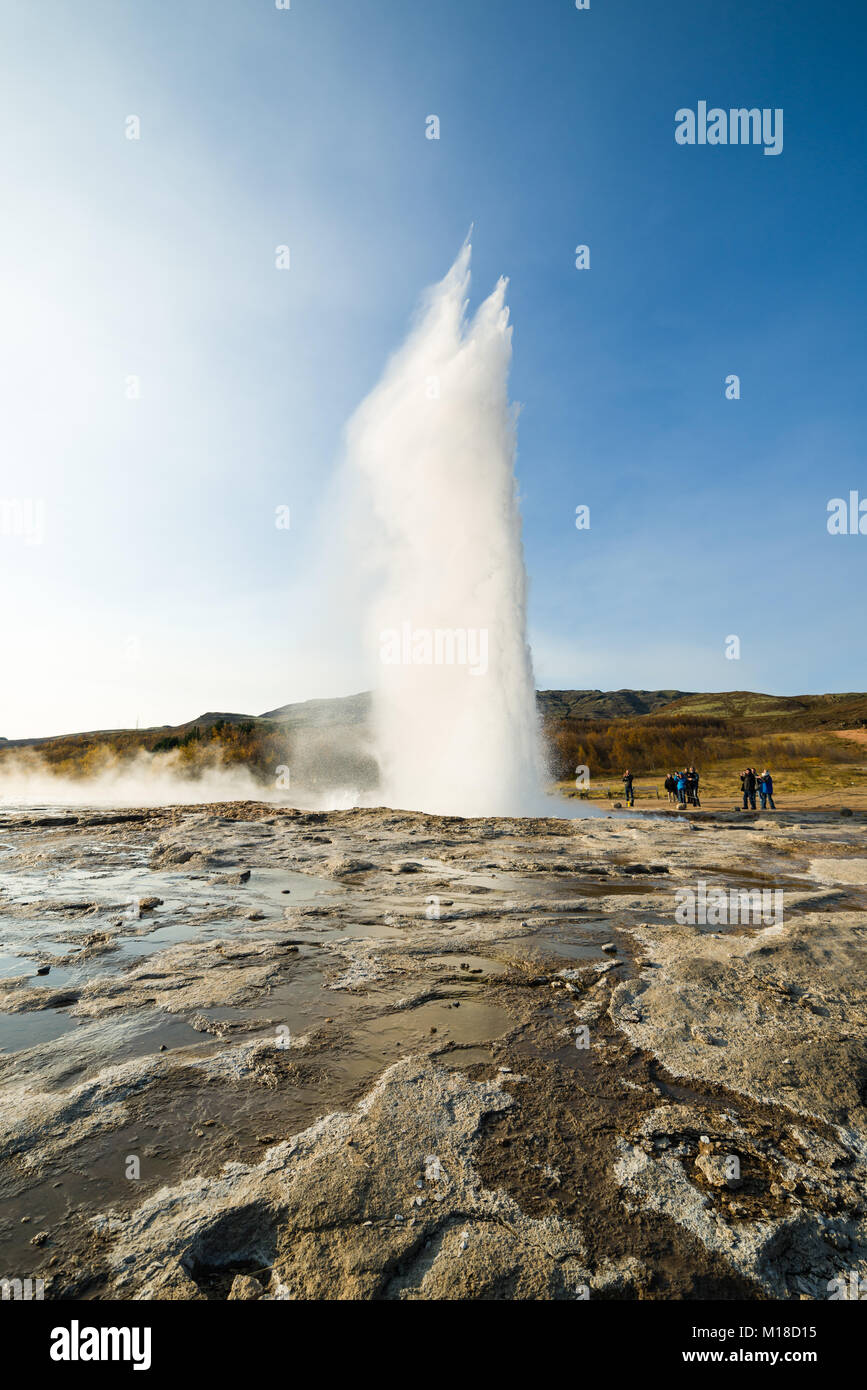 Tourists stand and watch Strokkur geyser erupting water due to geothermal activity on a sunny Autumn day, southwestern Iceland Stock Photo
