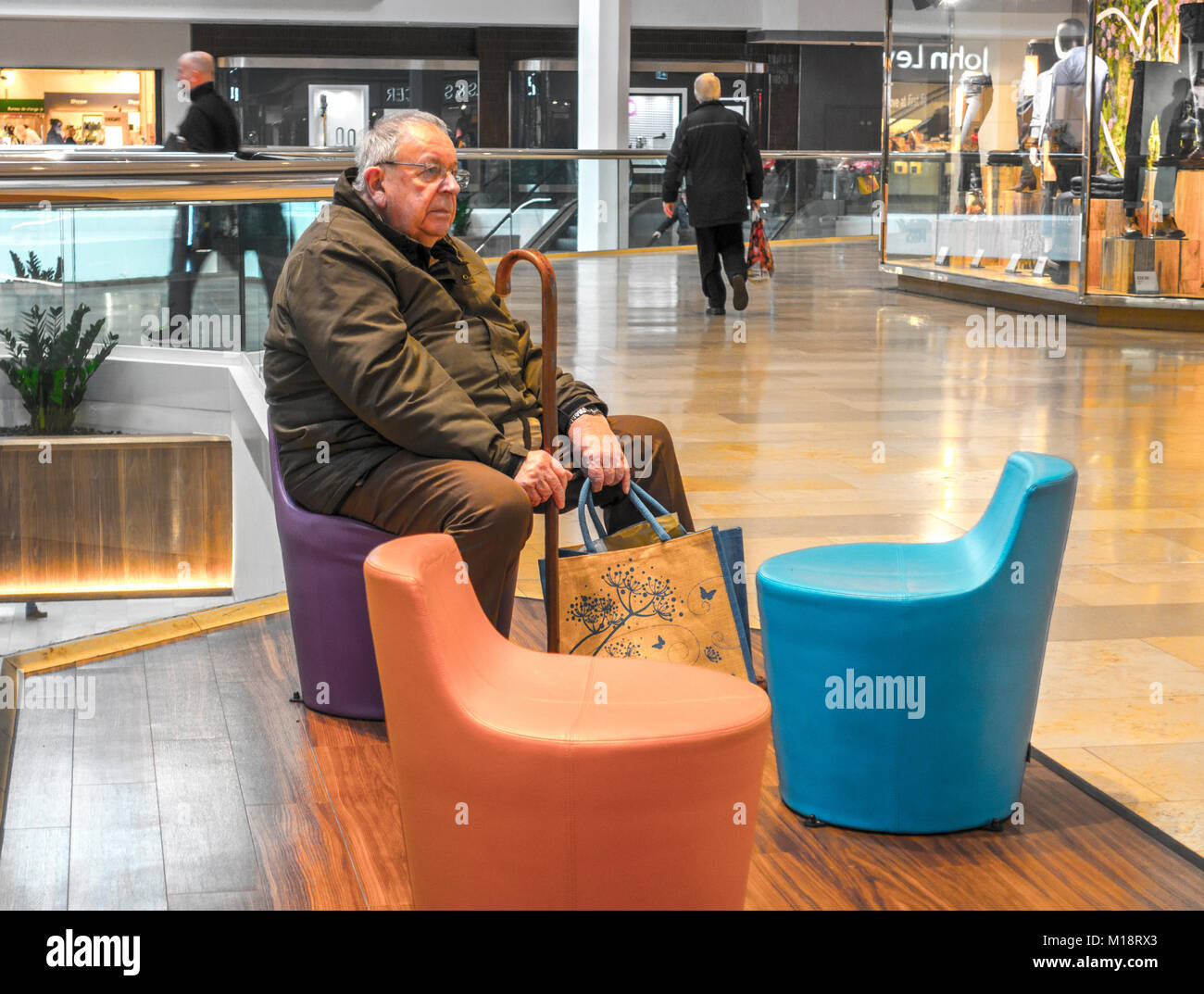 Old man sitting down, holding a walking stick and some carrier bags in the Queensgate shopping centre, Peterborough city, Cambridgeshire, England, UK. Stock Photo