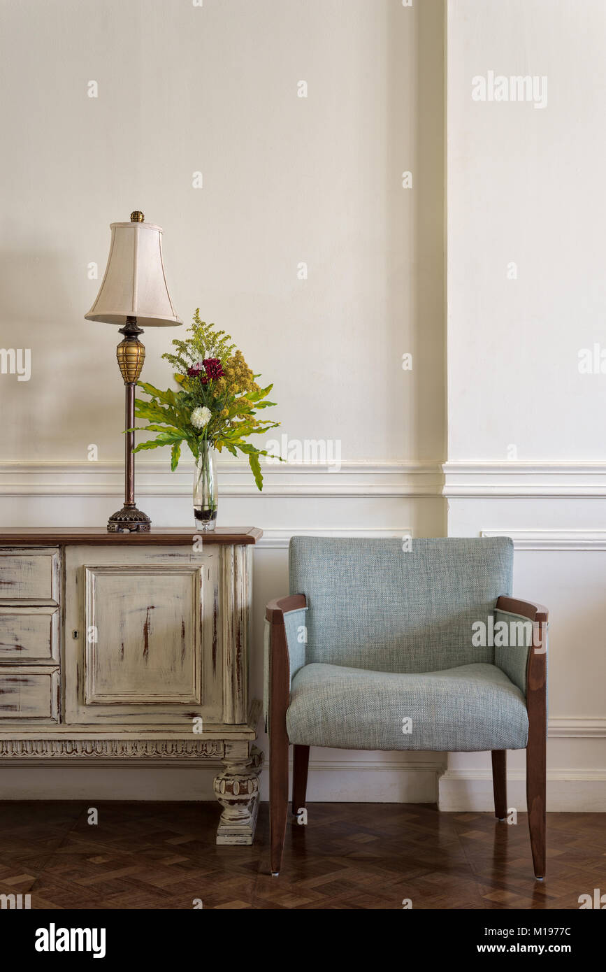 Interior shot of light blue armchair and cream vintage sidebar with table lamp and flowers planter on off white wall and wooden floor Stock Photo
