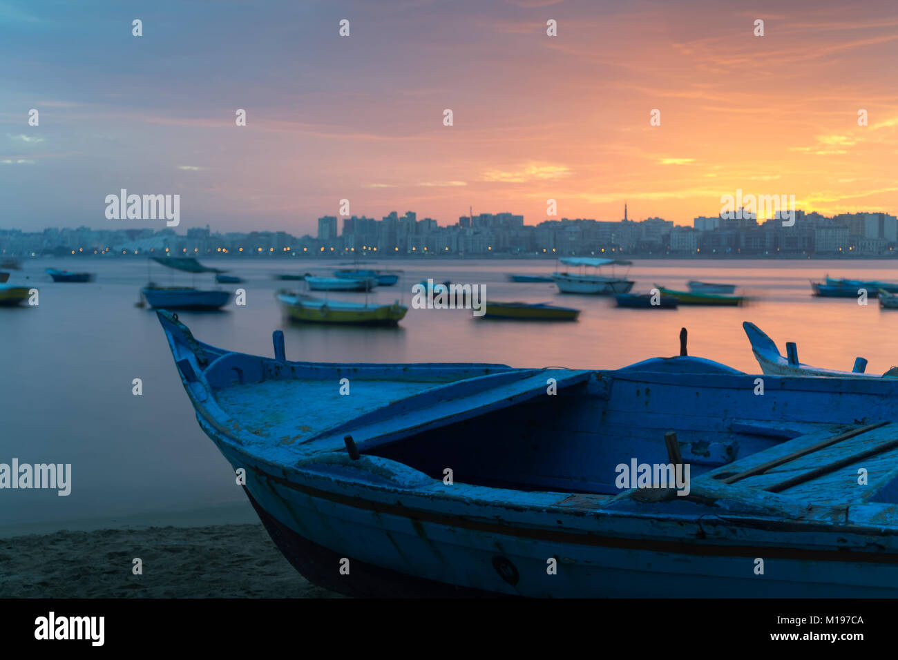 Turquoise blue fishing boat on the beach at sunrise with Alexandria skyline in far distance and colorful sky at sunrise Stock Photo