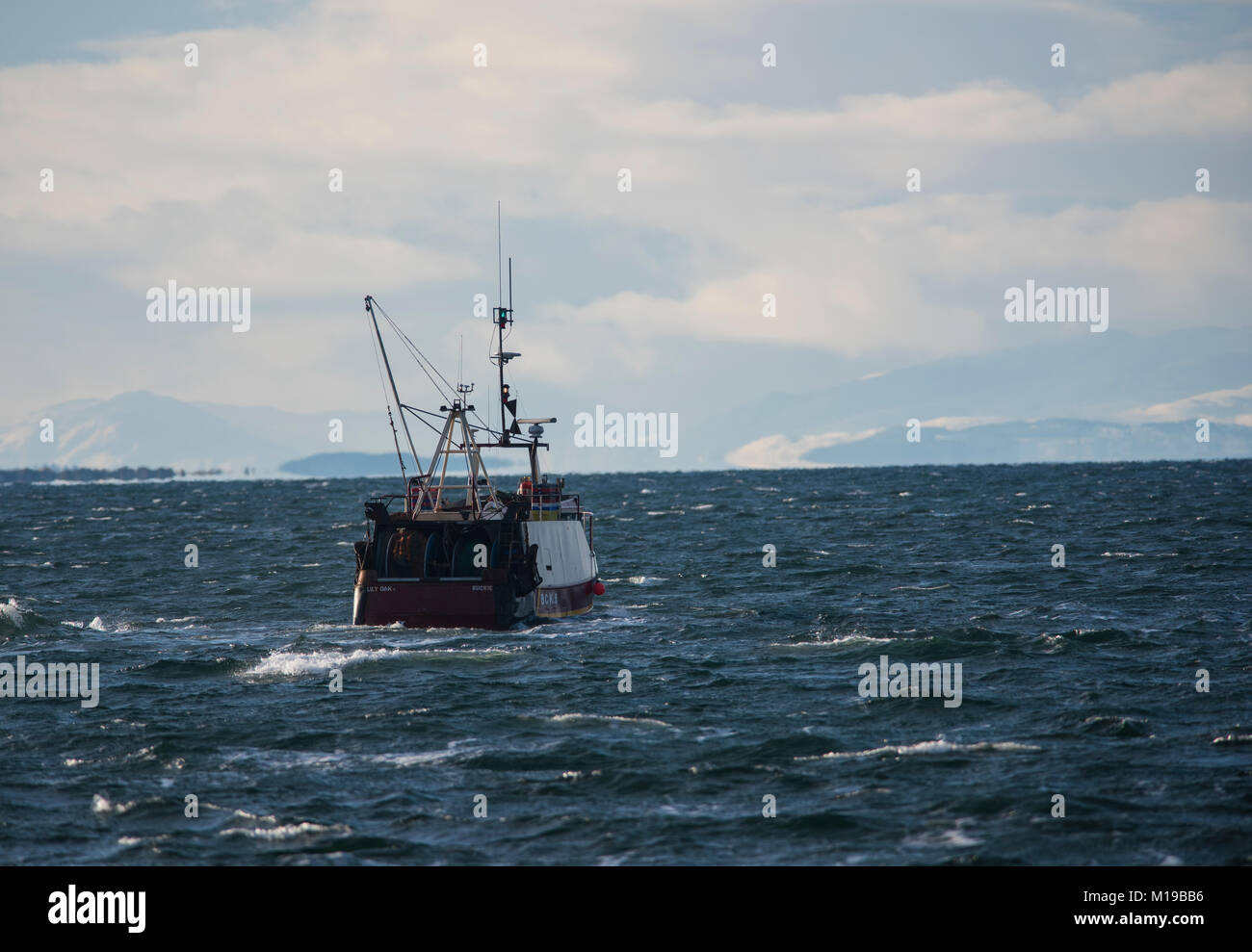 Fishing trawler leaving Burghead Harbour in the North of Scotland in winter with snow covered mountains in the background. Stock Photo