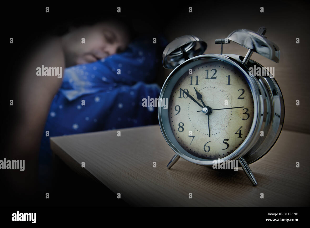 a man sleeping on the bed beside the alarm clock on table, in night. Stock Photo