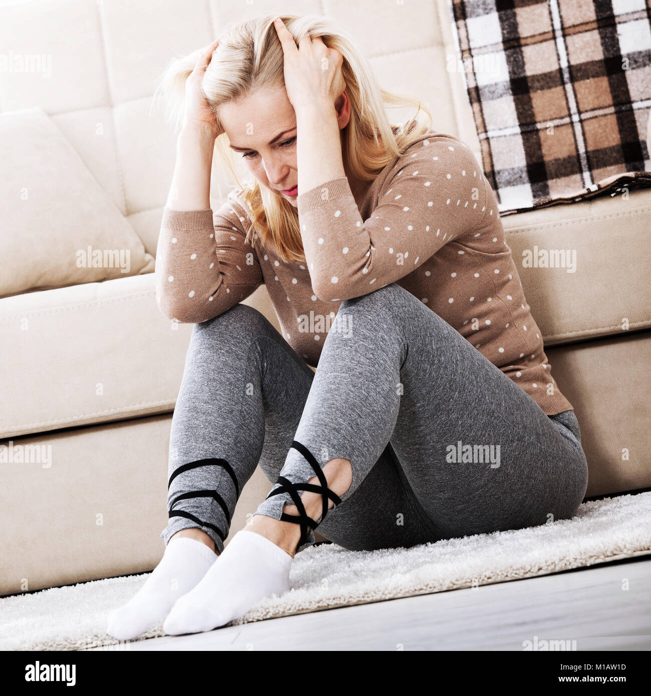 Depression. Middle aged barefoot woman sitting at the floor embracing her knees, near sofa at home, her head down, bored, troubled with domestic viole Stock Photo