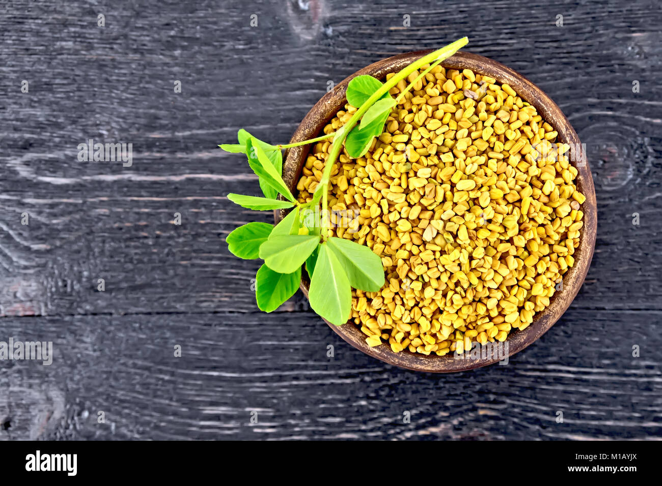 Fenugreek seeds in a clay bowl with green leaves on a wooden plank background Stock Photo