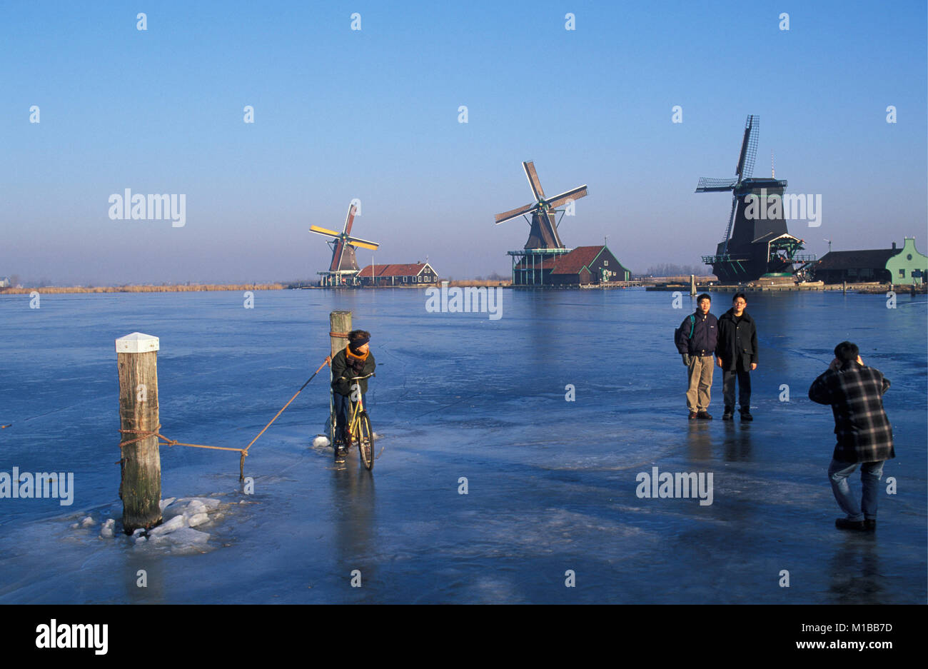 The Netherlands. Zaanse Schans. Historical windmill complex. Winter. Frozen river called De Zaan. Cyclist looking at Japanese tourists taking a pictur Stock Photo