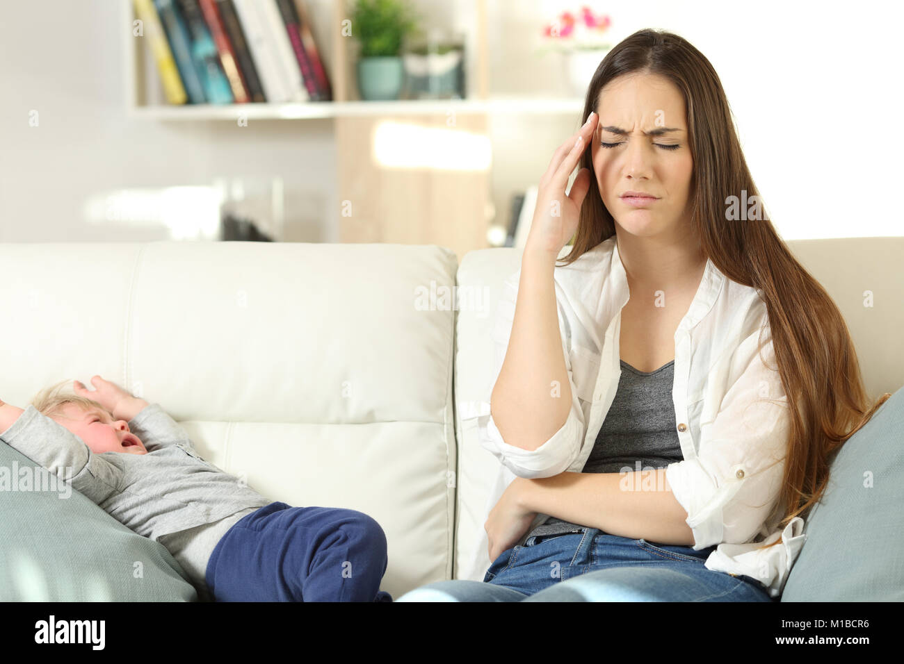 Front view portrait of an annoyed mother with her baby son crying sitting on a couch in the living room at home Stock Photo