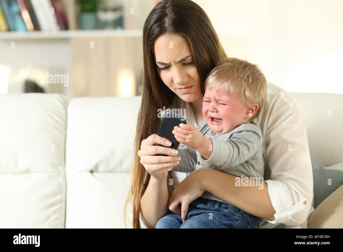 Baby having a tantrum and fighting with his mother for a smart phone sitting on a couch in the living room at home Stock Photo