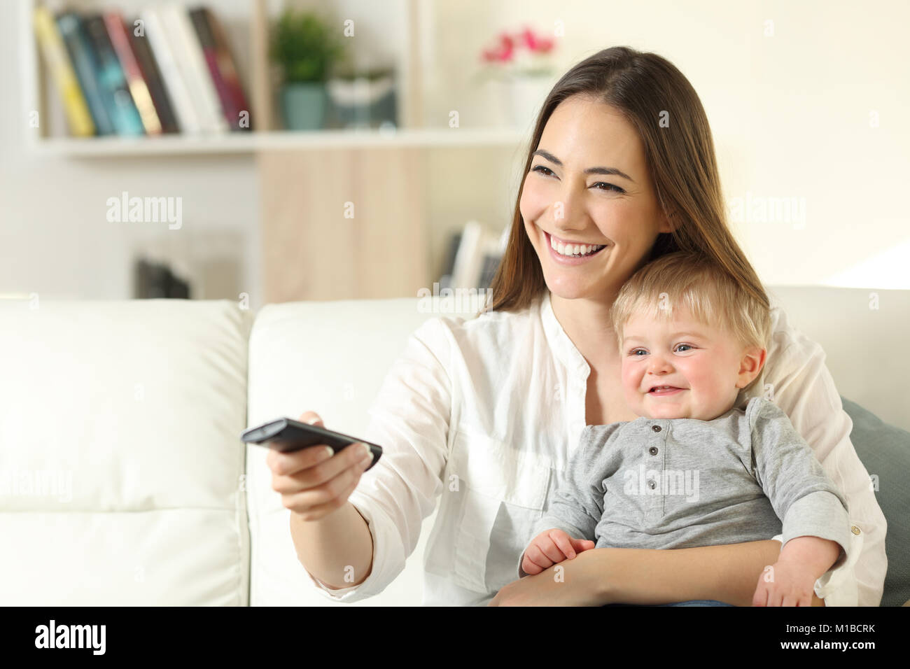 Happy mother and baby watching tv sitting on a couch in the living room at home Stock Photo