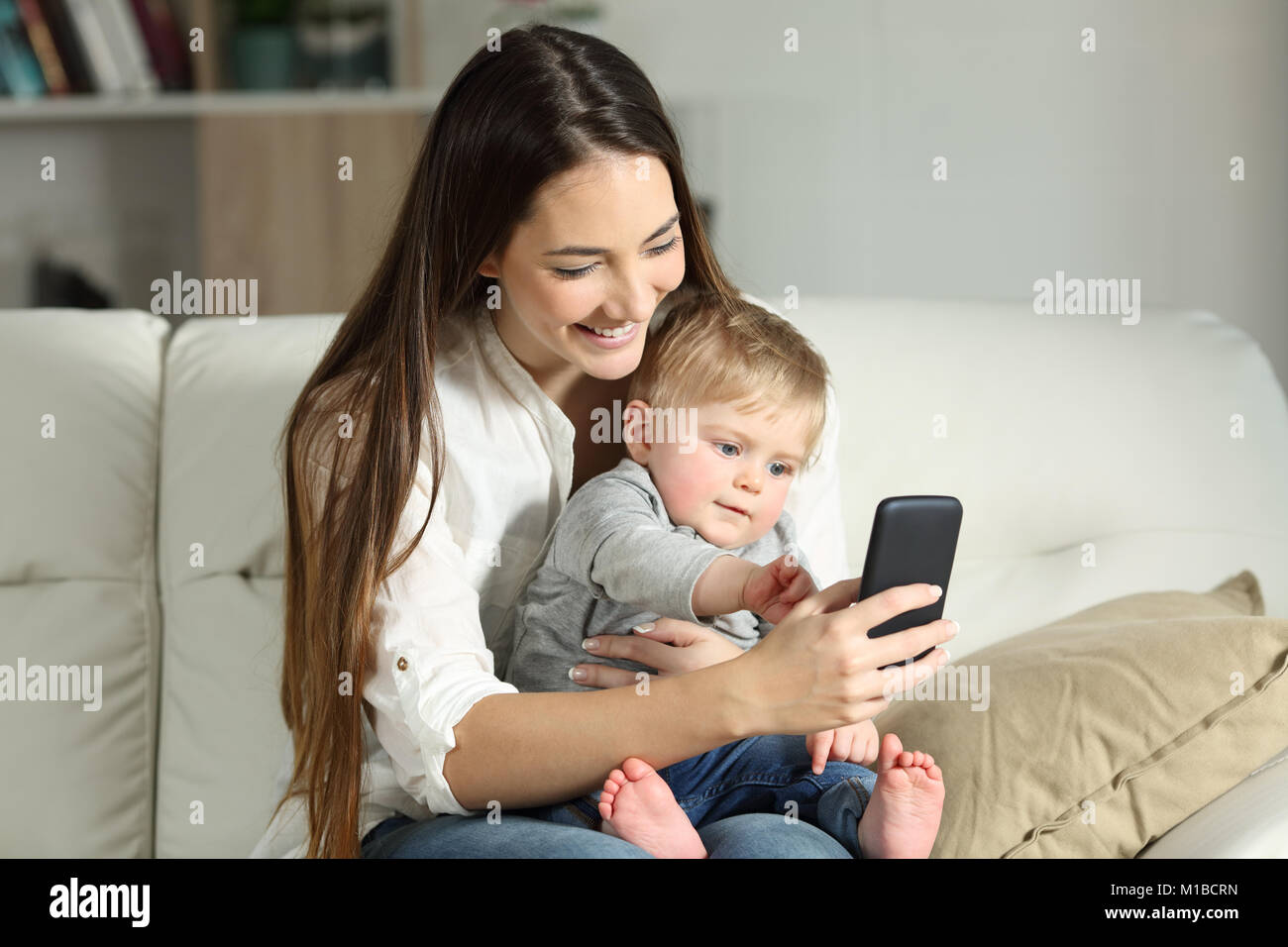 Mother and baby playing with a smart phone sitting on a couch in the living room at home Stock Photo