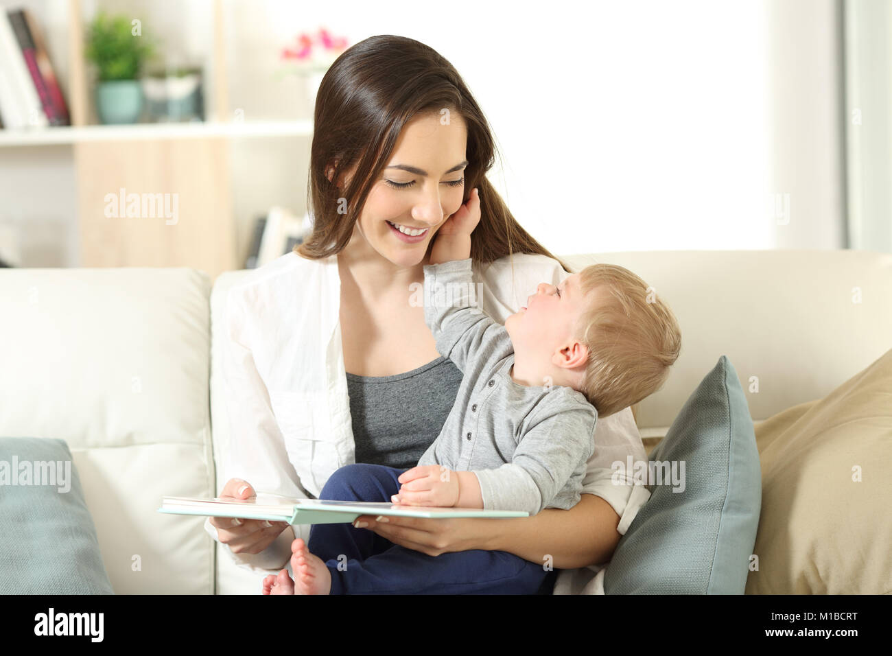 Mother and son looking each other holding a book sitting on a couch in the living room at home Stock Photo