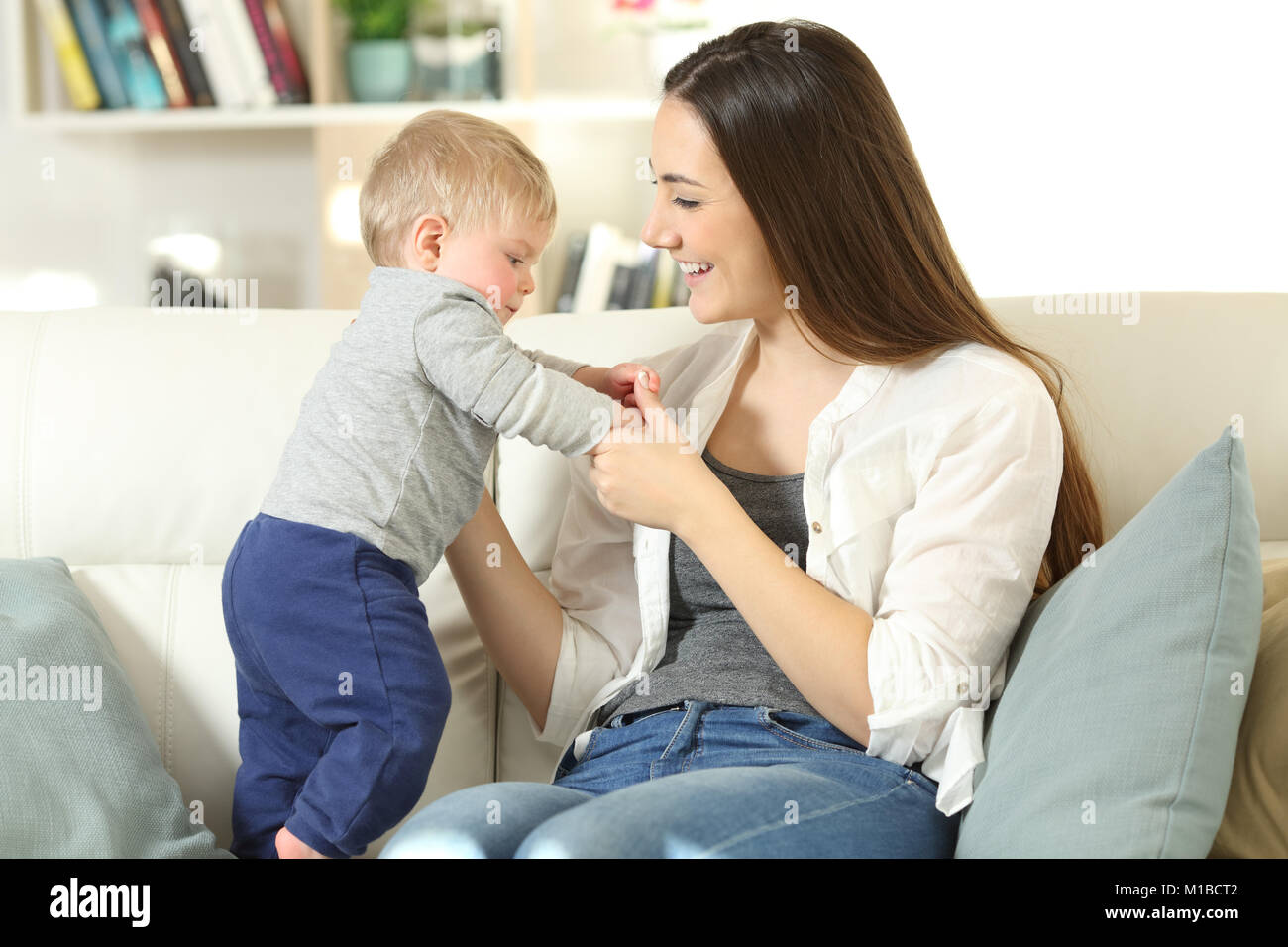 Proud mother helping to her baby to stand on a couch in the living room at home Stock Photo