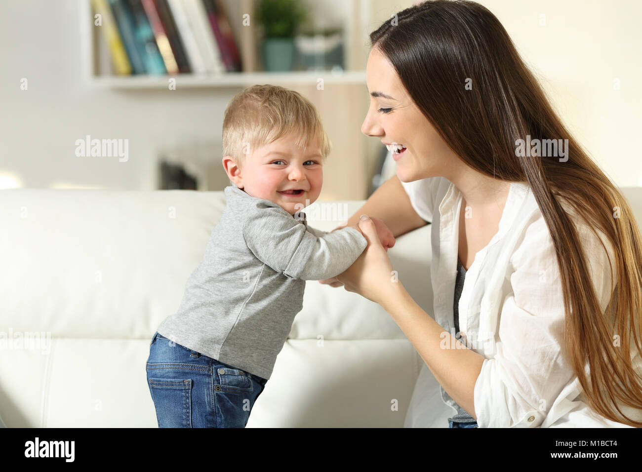 Mother helping to stand to her son who looks at you on a couch in the living room at home Stock Photo