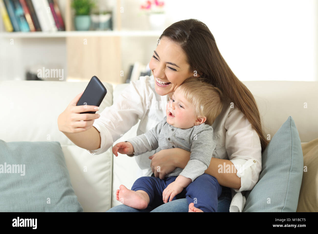 Mother taking a selfie with her baby son sitting on a couch in the living room at home Stock Photo