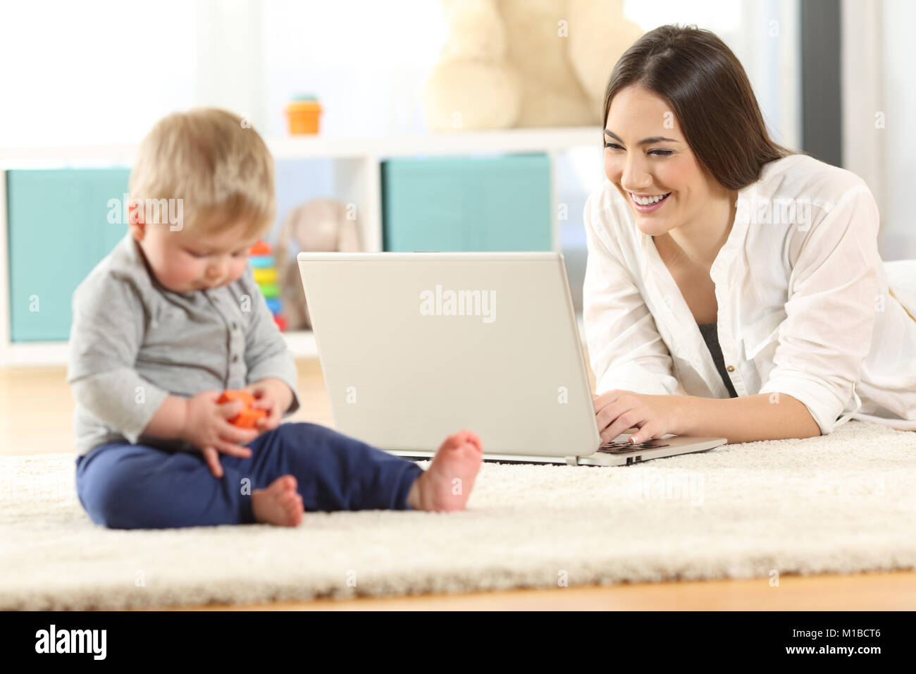 Mother working with a laptop and baby playing with toys on the floor at home Stock Photo
